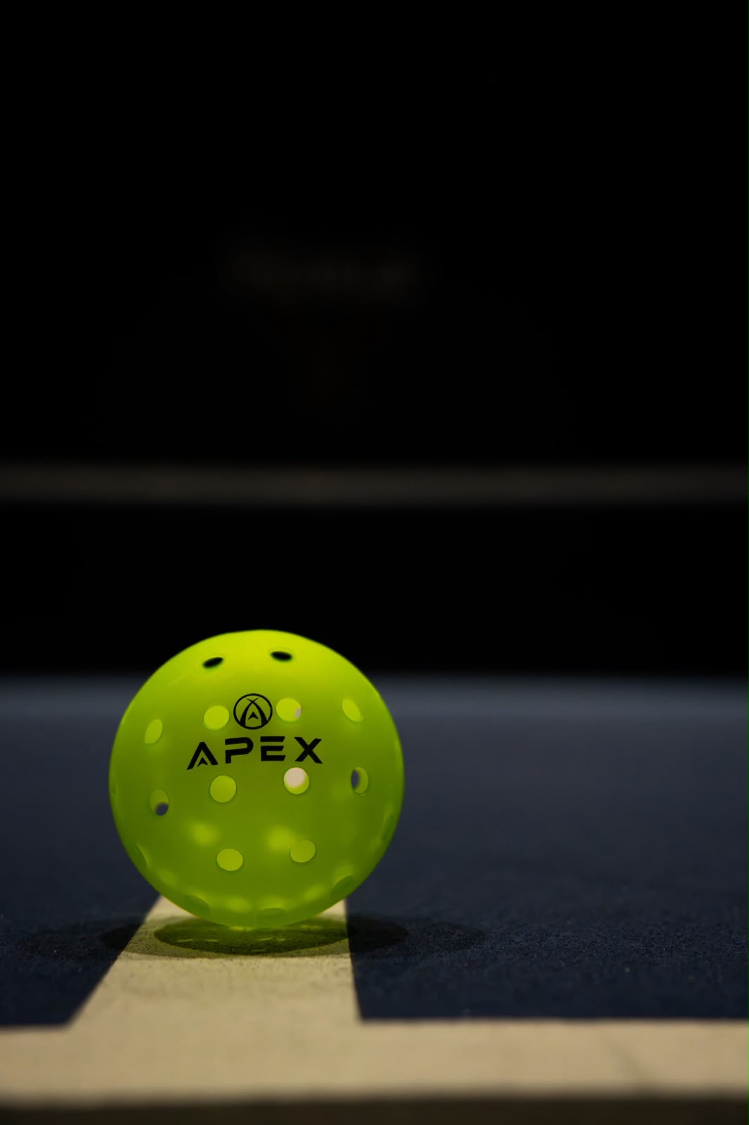 A bright neon yellow-green Apex ball resting on a dark blue and beige putting green surface against a dark background.