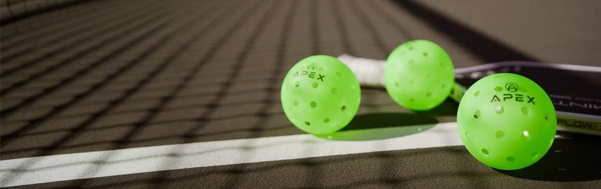 Three neon green Apex pickleball paddles are lined up on a dark tennis court with a white baseline, casting a long shadow.