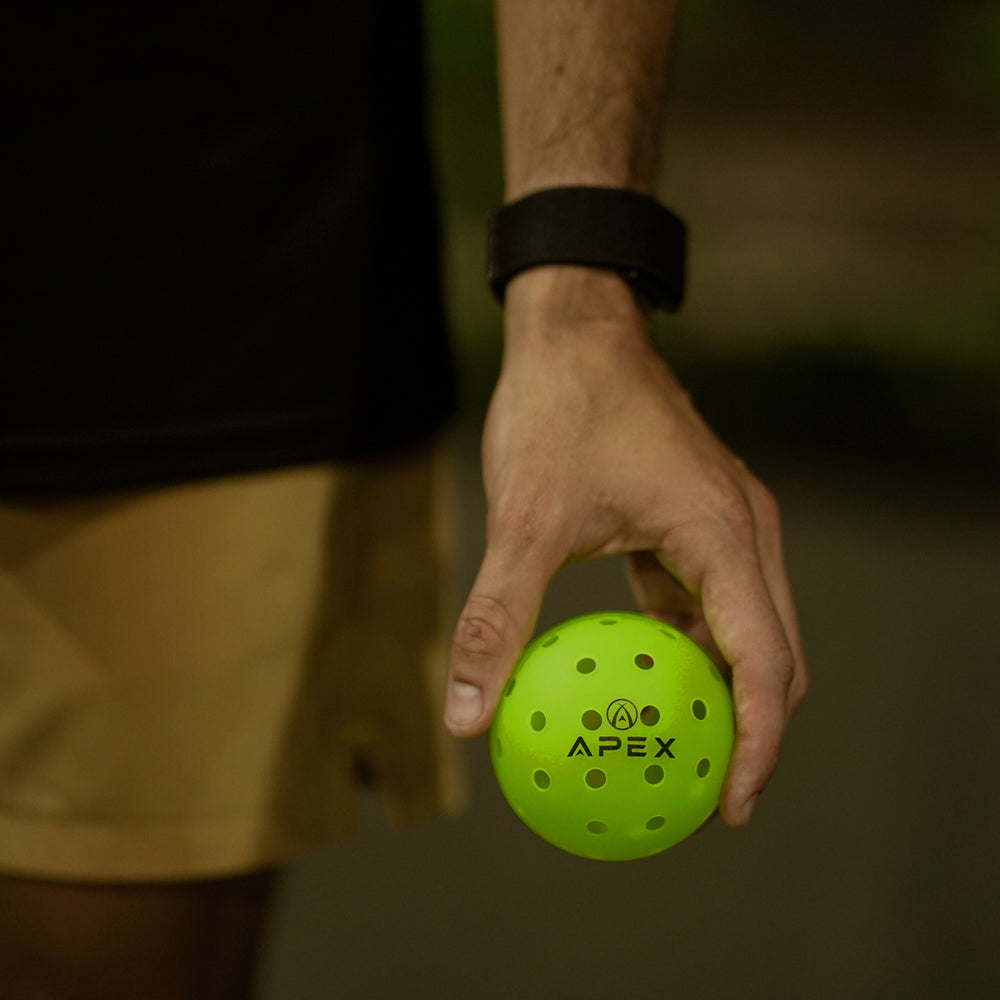 Player crouching on court, holding APEX pickleballs beside her shoe